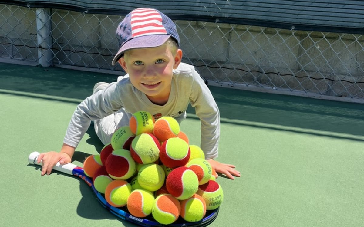 A young child on court behind a pyramid of red and orange junior balls, racquet in hand